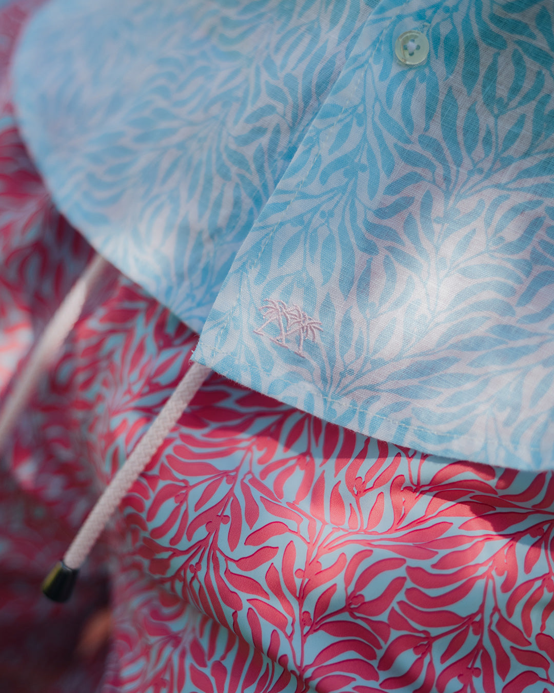 Close up of quality holiday clothing designed to last from Pink House showing a blue Sealeaf patterned linen shirt with matching sustainable swim shorts in coral pink and blue 