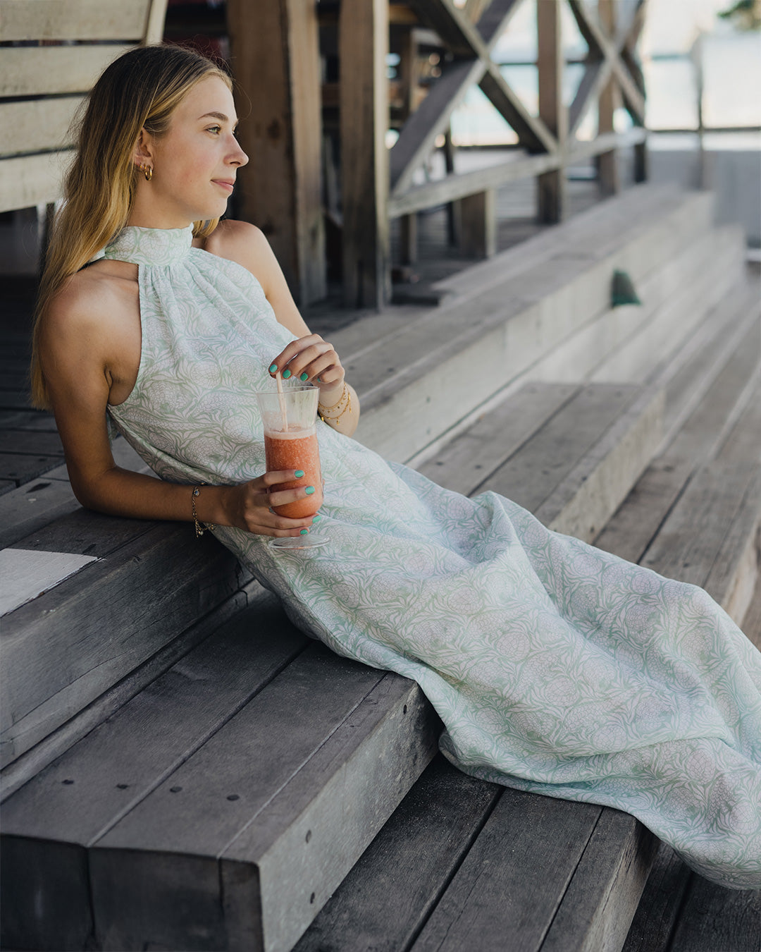 Woman wearing a long linen Trinny dress in green Pangolin design sitting on the steps with a rum punch at Basil's Bar, Mustique