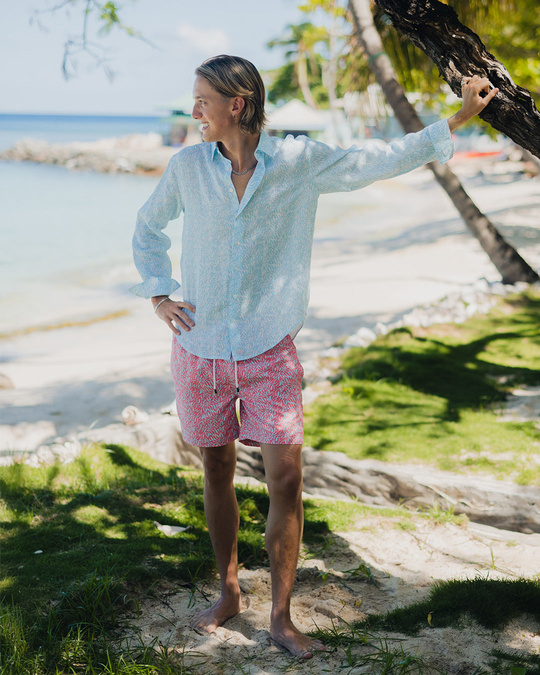 Man leaning on a tree on holiday wearing a blue Sealeaf patterned linen shirt with matching sustainable swim shorts in coral pink and blue 