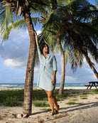 Woman wearing a Mustique Island vacation classic linen dress in light blue Sealeaf print, leaning against a palm tree on Lagoon Bay