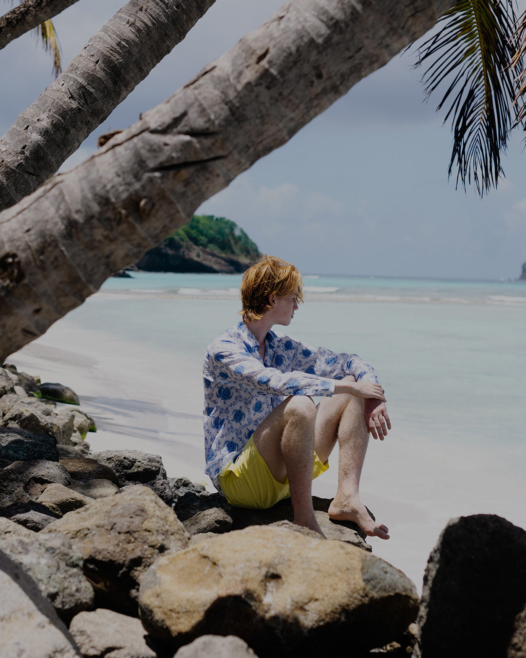 Man sitting on a tropical beach on vacation in a linen shirt with blue Turtle pattern and bright yellow swim shorts