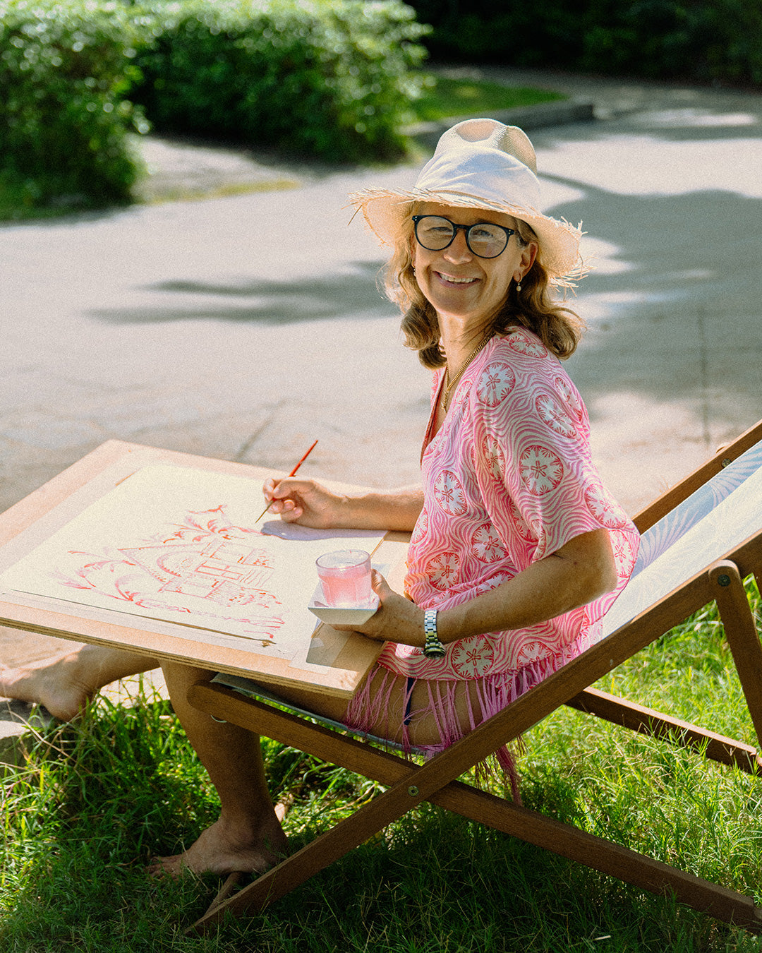 Designer Lotty wears pure silk poncho in coral pink Sand Dollar print whilst sitting painting outside the Pink House boutique shop on Mustique Island