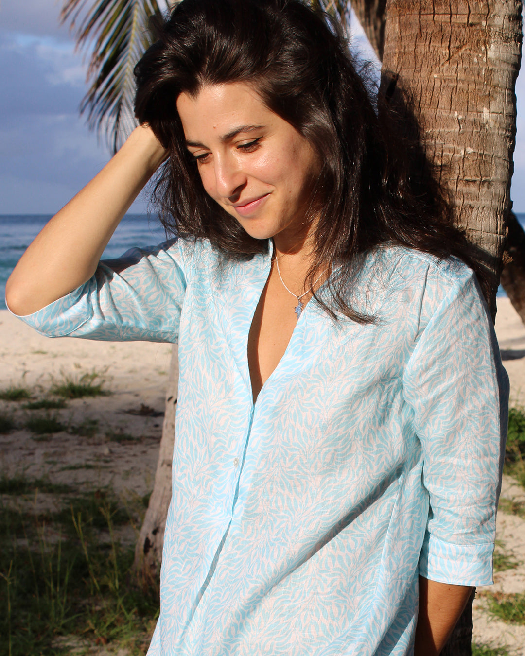 Woman leaning against a palm tree in a chic linen beach dress in blue Sealeaf design Lagoon Bay, Mustique Island