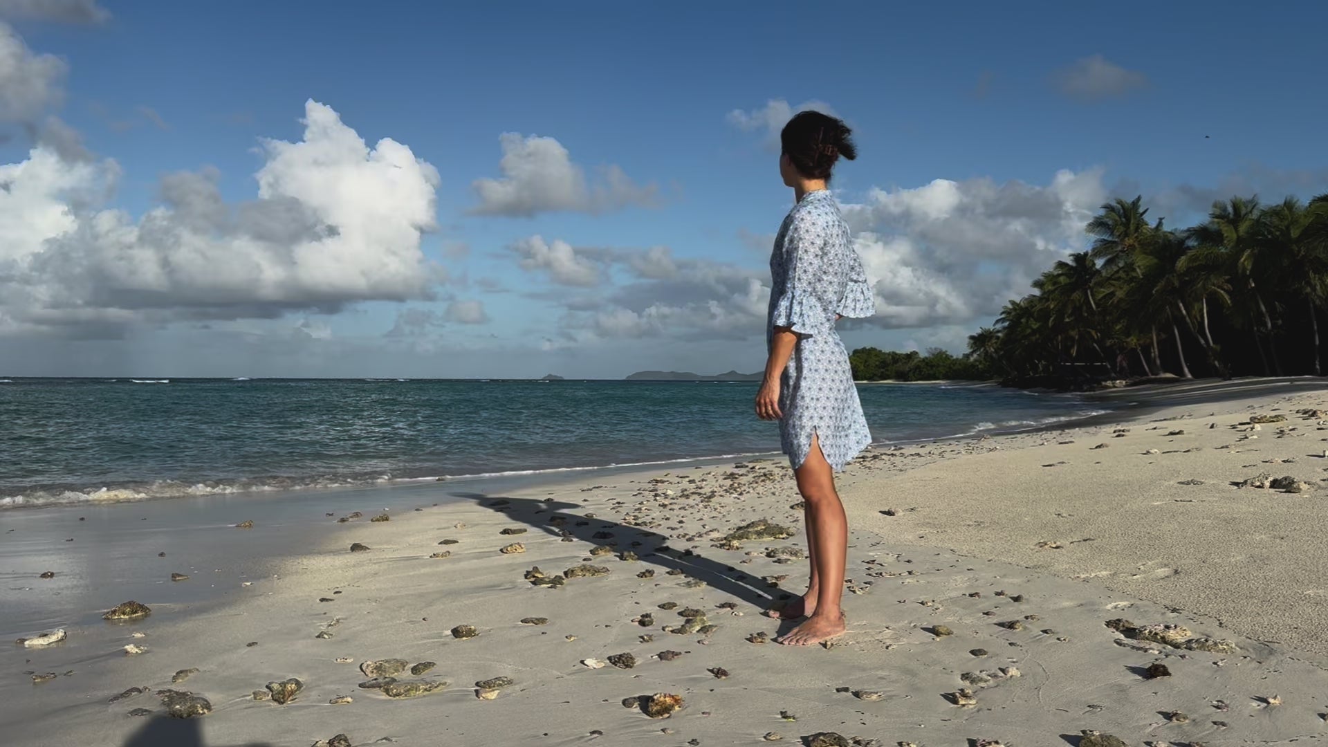 Woman styling a Decima linen beach dress with gathered 3/4 length sleeves in blue Sand dollar print, looking out to sea from Lagoon Bay, Mustque Island