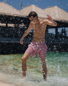 Man splashing in tropical water by Basil's Bar wearing sustainable swim shorts in coral pink and blue Sealeaf pattern